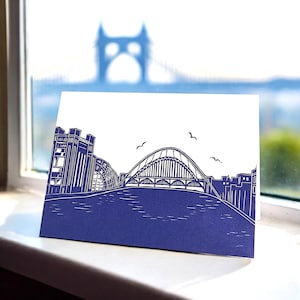 May include: A white card with a blue illustration of a bridge and buildings. The card is on a white windowsill.
