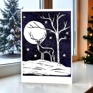 Könnte beinhalten: Ein Schwarz-Weiß-Linolschnitt eines Hirsches, der auf einem schneebedeckten Hügel steht, mit einem Vollmond und einem kahlen Baum im Hintergrund. Der Druck ist vor einem dunkelblauen Hintergrund mit weißen Sternen angeordnet. Die Karte wird auf einer weißen Oberfläche angezeigt.