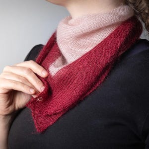 May include: A knitted scarf in a triangular shape, featuring a gradient of colors. The scarf transitions from a soft pink at the top to a deep red at the bottom. The texture of the knit is visible, suggesting a handmade item.