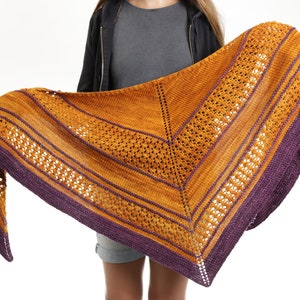 May include: A handmade shawl in shades of gold and purple. The shawl features a geometric design with openwork and striped sections. The edges are finished with a deep purple border. The shawl is held up, showcasing its triangular shape and intricate knit pattern.