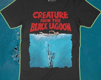 May include: A black short-sleeve t-shirt featuring the red text Creature from the Black Lagoon above a graphic of a person swimming on the water surface with a creature reaching up from the depths below. The shirt includes a Graveyard Classics label.