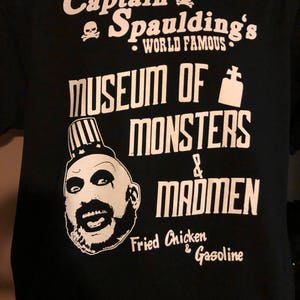 Puede incluir: Camiseta negra con texto blanco que dice "Captain Spaulding's World Famous Museum of Monsters & Madmen Fried Chicken & Gasoline". Un gráfico de calavera y huesos cruzados está por encima del texto. Una imagen de dibujos animados de un hombre con un sombrero a rayas y una gran sonrisa está debajo del texto.
