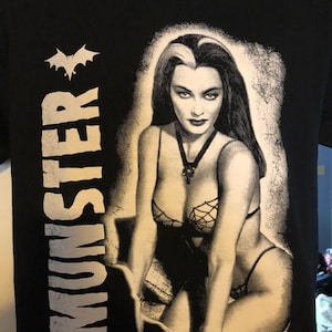 May include: Black t-shirt with a white graphic of a woman with black hair and a white streak in her hair. The graphic is in a distressed style and the woman is wearing a black and white lingerie-style outfit. The text "MUNSTER" is printed on the left side of the shirt in white.