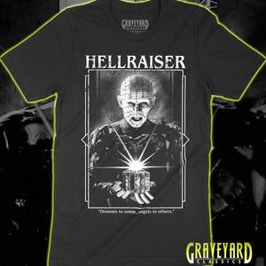 Puede incluir: Una camiseta negra de manga corta con un estampado gráfico blanco de Pinhead de Hellraiser sosteniendo una caja de rompecabezas. El texto HELLRAISER aparece sobre el personaje y la cita Demons to some, angels to others está impresa debajo.