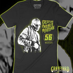 Pode incluir: Uma t-shirt preta de manga curta com um gráfico de uma múmia em branco com um brilho verde-lima. O texto diz "CREATURE DOUBLE FEATURE", "56" e "WLVI-TV" em verde-lima. A etiqueta interior do pescoço mostra "GRAVEYARD CLASSICS" e o tamanho M.