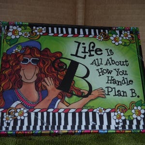 May include: A colorful framed art print with a cartoon woman with red hair and sunglasses. The text on the print reads "Life is all about how you handle Plan B."