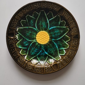 May include: A brown ceramic ashtray with a green and blue flower design. The flower has a yellow center with a textured pattern.