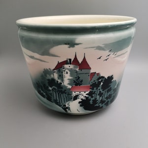 May include: A white ceramic planter with a green, pink, and black scene of a castle in a forest. The castle has red roofs and a bridge.