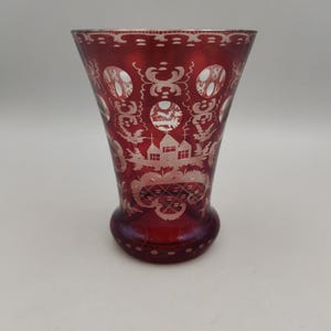 May include: A red glass vase with a white etched design. The design features a house and other geometric patterns.