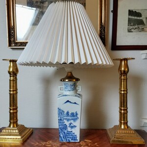 May include: A white pleated lampshade sits atop a blue and white porcelain vase with a landscape scene. The vase is on a wooden table with two gold candle holders.