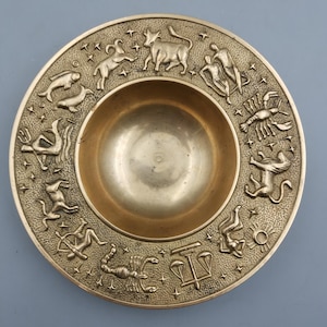 Puede incluir: Un plato redondo de latón antiguo con un cuenco central y un borde exterior en relieve. El borde presenta símbolos del zodíaco y estrellas en relieve. El plato tiene un brillo metálico cálido y está sobre un fondo gris apagado.
