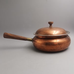 Pode incluir: Uma panela de cobre com cabo e tampa de madeira. A panela tem um corpo arredondado e uma borda prateada. A tampa também é de cobre com um botão de madeira. A alça é feita de madeira e é presa à panela com uma conexão de latão.