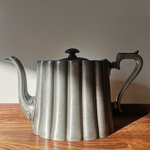 May include: A vintage silver teapot with a fluted body, a curved spout, and a handle. The lid is topped with a black knob. The teapot is displayed on a wooden surface.