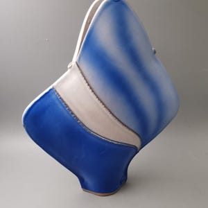 Puede incluir: Un jarrón de cerámica con una forma geométrica única. El jarrón presenta una llamativa combinación de colores azul y blanco, con una superficie texturizada. El diseño incluye una banda blanca central y rayas azules, creando una estética moderna.