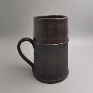 Peut inclure: Une tasse en céramique brun foncé avec une finition texturée. La tasse a une large poignée arrondie et une lèvre légèrement évasée.