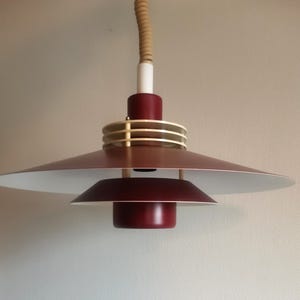 Puede incluir: Una moderna lámpara colgante con un diseño en capas. La lámpara presenta una pantalla de color burdeos con interior blanco, un anillo dorado y un cable beige. La lámpara está suspendida del techo.