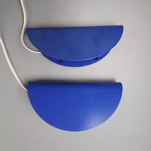 Può includere: Due applique da parete semicircolari blu cobalto con cavi bianchi. Le applique sono montate su una superficie grigio chiaro. L'applique superiore è posizionata sopra quella inferiore.