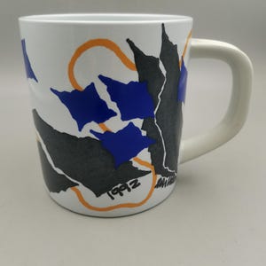 May include: White ceramic mug with a curved handle. The mug features a design of blue, black, and orange abstract shapes. The year "1992" is printed on the side of the mug.