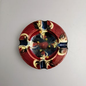May include: A round, ceramic ashtray with a red rim and a dark blue center. The ashtray features a decorative floral design in yellow, blue, and red. It has three notches for resting cigarettes.