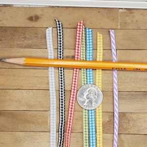 May include: Seven different colored gingham ribbons with a pencil and a US quarter on a wooden surface. The ribbons are black and white, red and white, yellow and white, green and white, blue and white, and purple and white.