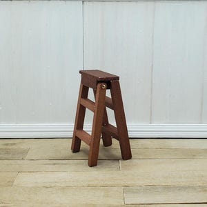 May include: A miniature wooden step stool with a dark brown finish. The small ladder has two steps and a top platform, connected by a brass hinge. The stool is set against a white wall and light wood floor.