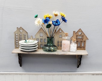 May include: A wooden shelf with a cityscape design, featuring a vase with blue and yellow flowers, three small glass jars, and a stack of white plates.