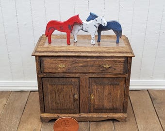 May include: A miniature wooden cabinet with a dark brown finish, one drawer, and two doors with small gold-toned handles. Three small horse figurines in red, white, and dark blue are placed on top. A copper penny is positioned in the foreground.
