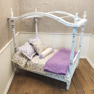 May include: A miniature four-poster bed with a distressed white painted finish. The bed is dressed with floral patterned bedding in shades of purple and green, two square pillows, a small cylindrical bolster pillow, and a fuzzy purple blanket.