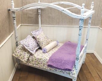 May include: A miniature four-poster bed with a distressed white painted finish. The bed is dressed with floral patterned bedding in shades of purple and green, two square pillows, a small cylindrical bolster pillow, and a fuzzy purple blanket.