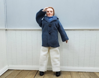 May include: A doll wearing a navy blue coat, a light blue sweater, and white pants. The doll is also wearing black shoes. The doll is saluting with their right hand. The background is a light gray wall and white wainscoting.