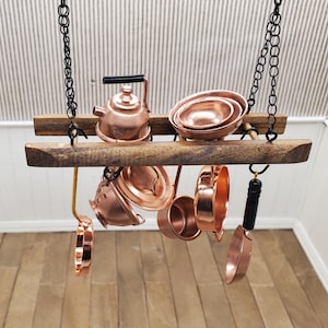 May include: Miniature copper kitchenware set hanging from a wooden rack. The set includes a kettle, pots, pans, and a ladle. The copper items are suspended by black chains, creating a rustic and charming display. The rack is made of dark wood.
