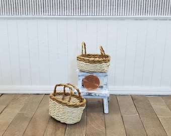 May include: Two miniature woven baskets with handles. One basket sits on a small, white, distressed stool, with a penny on top. The other basket is on the floor. The baskets are beige and brown.