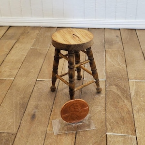 May include: A miniature wooden stool with a round seat and four legs. The stool is made of light brown wood and has a classic design. A penny is placed next to the stool for scale.