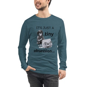 Puede incluir: Una camiseta de manga larga de color azul verdoso con una imagen en blanco y negro de una persona sosteniendo una casa de muñecas. El texto "It's just a tiny obsession..." está impreso en la camiseta.