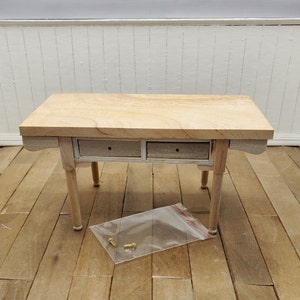 May include: A light wood table with two drawers and four legs. The table is on a wood floor. There are four small gold screws in a plastic bag on the floor.