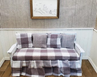 May include: A miniature white sofa with a checkered pattern in shades of white and brown. The sofa has four matching pillows and a skirted base. A framed landscape art piece hangs above the sofa on a striped wall.