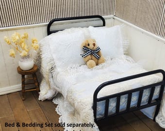 May include: A miniature black metal bed frame featuring white lace bedding, two lace pillows, and a small tan teddy bear wearing a black and white gingham bow. The text Bed & Bedding Set sold separately. Accessories not included. is visible on the image.