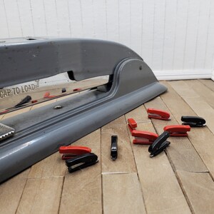 May include: A gray metal stapler with the text "CAP TO LOAD" on the front. The stapler is surrounded by eight miniature red and black staplers.