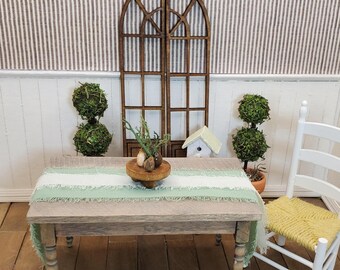 May include: A miniature rustic table with a green and white fringed runner, topped with a wooden tray holding decorative items. Two topiary trees flank the table, with a white chair and a decorative window in the background. A small birdhouse is also present.