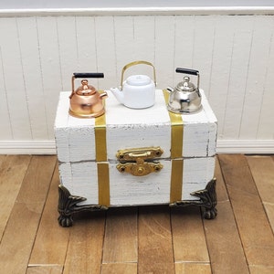 May include: Miniature tea kettles in copper, white, and silver sit atop a white chest with gold accents and ornate metal hardware. The chest has gold bands and decorative corner pieces, set on a wooden surface.