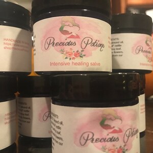 Puede incluir: Tres pequeños frascos de vidrio con tapas negras y etiquetas blancas. Las etiquetas tienen un fondo rosa con un diseño floral y el texto "Precious Potions" y "Intensive healing salve".