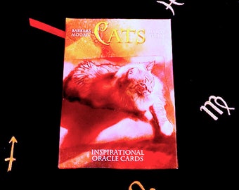 May include: A red and gold oracle card deck with a cat on the front. The card reads "Cats Inspirational Oracle Cards" with the authors' names Barbara Moore and Marco Tuzzini. The card is on a black background with gold zodiac symbols.