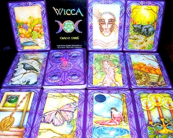 May include: A set of Wicca oracle cards with purple borders and colorful illustrations. The cards feature images of a crow, a butterfly, a tree, a pumpkin, a cat, a woman, and candles. The box for the cards has a holographic image of three moons and a star.