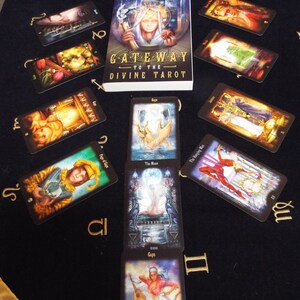 May include: Tarot card reading spread on a black velvet cloth with gold accents. The cards are from the Gateway to the Divine Tarot deck by Giro Marchetti. The cards are arranged in a line with the book in the center. The cards are The Page of Cups, The Moon, The Hanging Man, and The Eight of Cups.