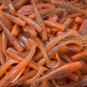 May include: Close-up of a pile of soft plastic fishing lures. The lures are a translucent orange color with glitter flecks throughout. Each lure has a paddle tail and a slender body, designed to mimic small fish.