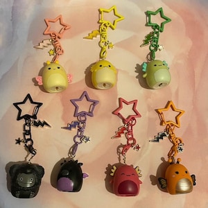 Op de afbeelding: Collectie kleurrijke sleutelhangers met verschillende stripfiguren. Elke sleutelhanger heeft een stervormige bovenkant, bliksemaccenten en een kleine bedel. De karakters zijn onder meer een gele kuiken, een groene axolotl en een zwart monster.