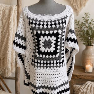 Op de afbeelding: Een witte, zwarte en grijze gehaakte poncho, tentoongesteld op een mannequin. De poncho heeft een vierkant patroon in het midden en is gemaakt van zacht, getextureerd garen. De tekst "WHEN YOU" is zichtbaar op de halslijn.