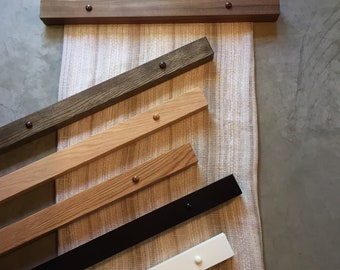May include: A collection of rectangular wooden poster hangers in various finishes including light wood, dark wood, stained wood, black, and white, each featuring two small circular knobs on the front, displayed over a textured beige fabric.