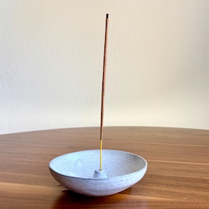 May include: A round, speckled white ceramic incense holder with a small central cone for holding a single incense stick. A thin, brown incense stick is inserted upright into the center of the holder, with a small dark tip at the very top.