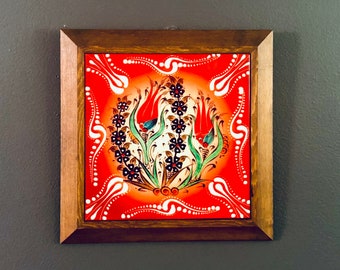May include: A framed ceramic tile with a red background and a floral design featuring three stylized tulips in shades of red, green, and blue. The tile is surrounded by a brown wooden frame.